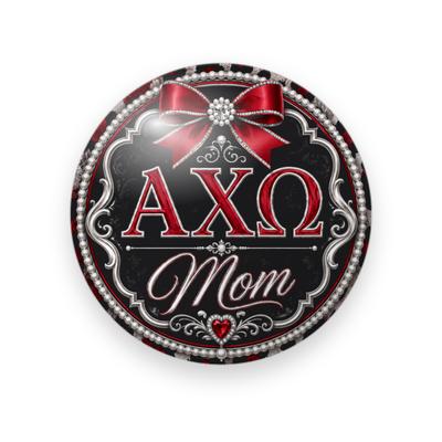 Alpha Chi Omega #045 button. Black background with a pearl and silver filigree frame, red Greek letters, red script “Mom” outlined in silver, a red bow and a jeweled heart.