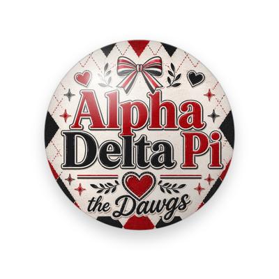 Alpha Delta Pi #066 button. Cream and black argyle background with the full name spelled out in red and black serif, a red bow, hearts and black script “the Dawgs.”