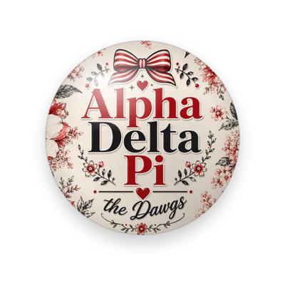 Alpha Delta Pi #081 button. Cream background with red and black florals, the full name spelled out in red and black serif, a striped red bow, a heart and black script “the Dawgs.”