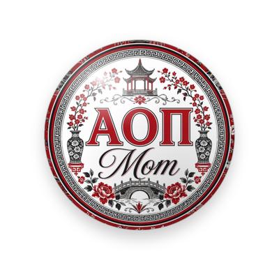 Alpha Omicron Pi #044 button. White toile with red and black pagodas, vases, a bridge and cherry blossoms inside a Greek key border, red Greek letters and black script “Mom.”