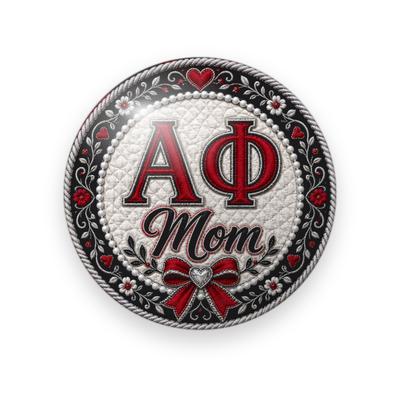 Alpha Phi #074 button. White quilted center with red Greek letters, red script “Mom”, and a red bow, framed by a black floral border with pearls and hearts.