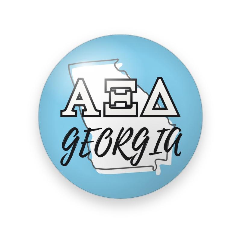 Alpha Xi Delta #046 button. Light blue background with a white Georgia state outline behind white block Greek letters outlined in black and black script “GEORGIA.”