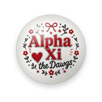 Alpha Xi Delta #053 button. White linen background with red stitched “Alpha Xi” lettering, a red heart, black script “the Dawgs”, a red bow, and red flowers and leaves.