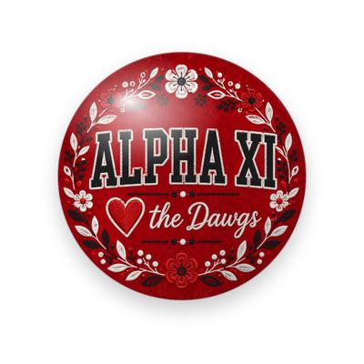 Alpha Xi Delta #054 button. Red floral background with black block “ALPHA XI” lettering outlined in white, a red heart outline, white script “the Dawgs”, and white flowers.